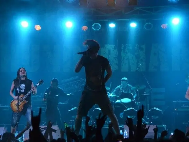 The image shows a group of people on stage at a concert, with some of them holding guitars and one...