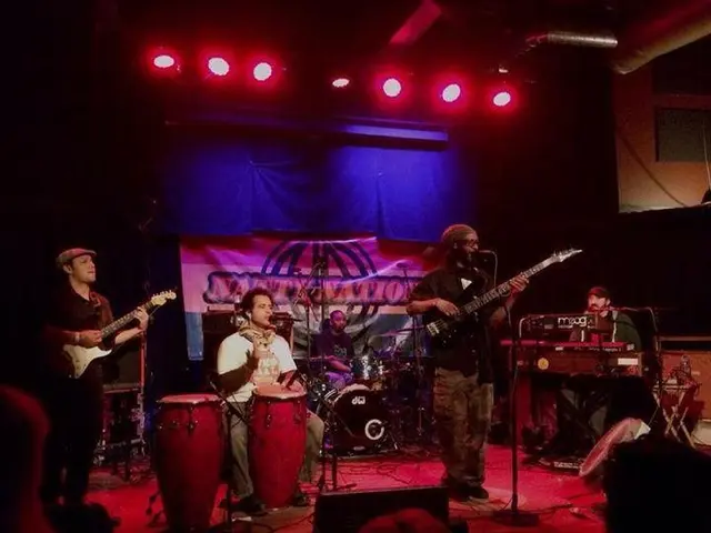 The image shows a group of people on stage playing musical instruments, with mics and stands set up...