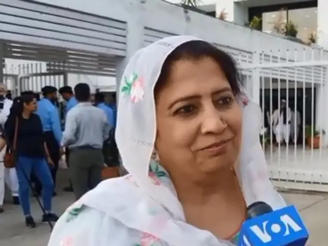 The image shows a woman in a white sari holding a microphone, surrounded by a group of people...