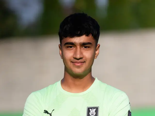 The image shows a young man in a green shirt standing on a soccer field with his arms crossed,...