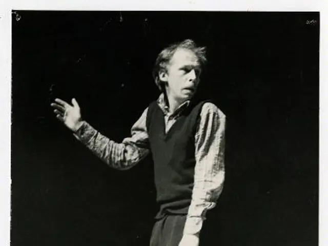 The image shows a man standing on a stage in a black and white photo. He is wearing a white shirt...
