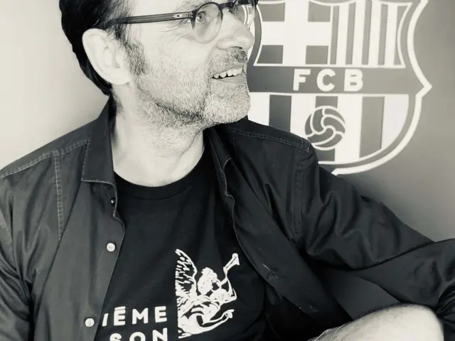 The image shows a man wearing glasses and a black shirt sitting in front of a wall with the FC...