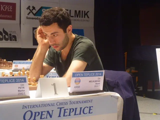 The image shows a man sitting at a table with a chess board in front of him, surrounded by chess...