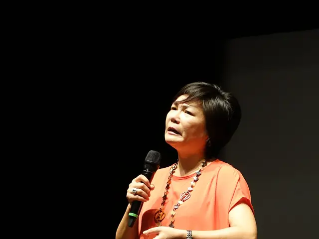 The image shows a woman standing on top of a stage, holding a microphone in her hand. She has a...