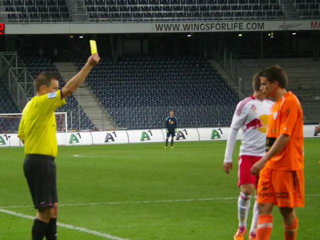 The image shows a referee showing a yellow card to a soccer player on the field surrounded by a...
