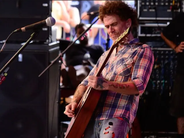 The image shows a man playing a guitar on a stage at a music festival. He is wearing a plaid shirt...