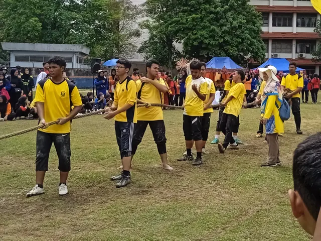 The image shows a group of people in yellow shirts playing tug of war on a grassy field. Some of...