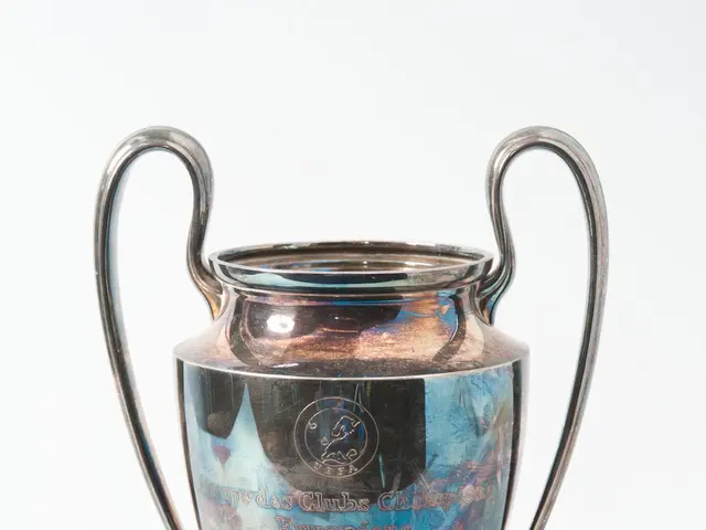 The image shows the UEFA Champions League Trophy, a steel cup with two handles and a blue and...
