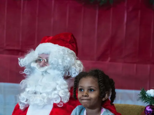 The image shows a little girl sitting on a couch next to Santa Claus, holding a book in her hands....