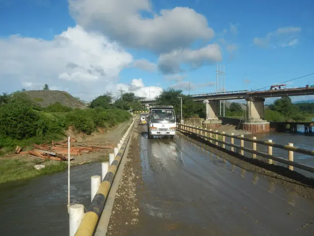The image shows a truck driving down a flooded road next to a bridge, with railings on either side....