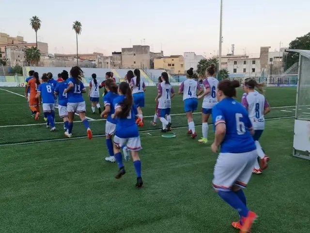 The image shows a group of women playing soccer on a field surrounded by buildings, trees, poles,...