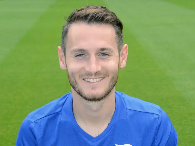 The image shows a man sitting on top of a soccer field, wearing a blue t-shirt and smiling. He...