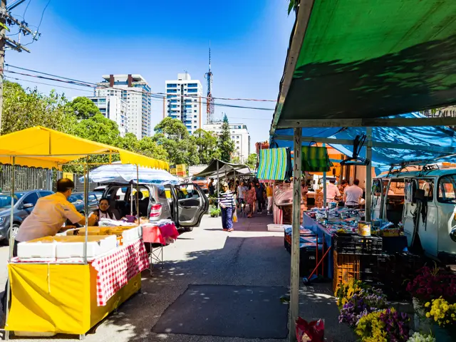 The image shows a bustling street market with people walking down the street next to parked cars....