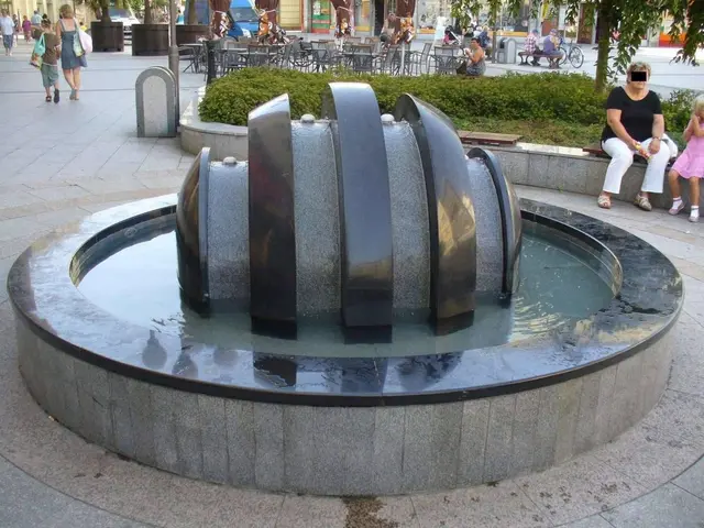 The image shows a fountain in the middle of a city square surrounded by people walking around it....
