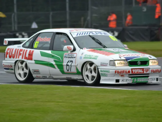 The image shows a white and green car driving on a race track, surrounded by grass on either side....