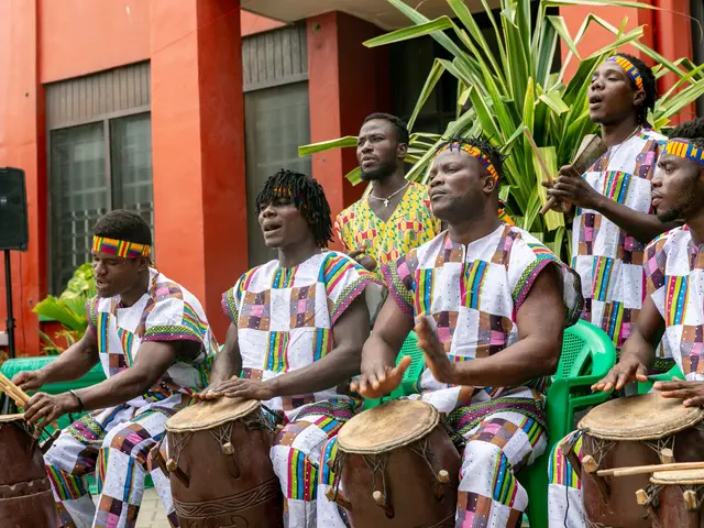 The image shows a group of men playing drums in front of a building, surrounded by chairs, plants,...