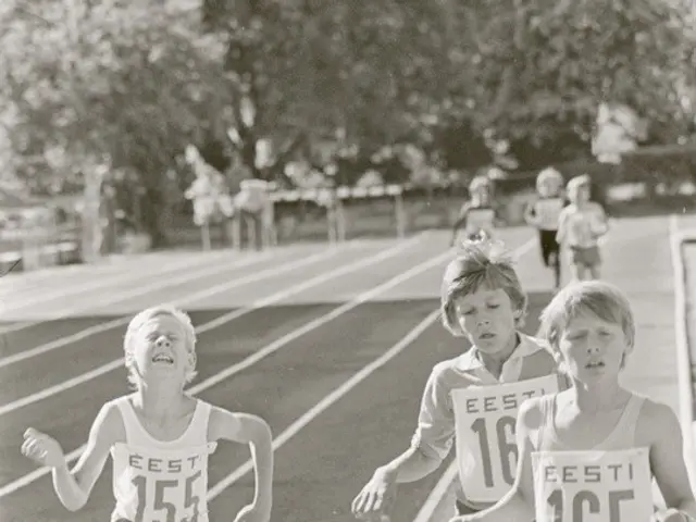The image shows a group of young boys running on a track surrounded by trees and other people in...