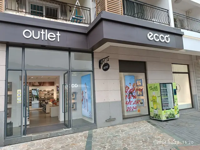 The image shows an outlet store in the middle of a city street, with glass walls and doors, posters...