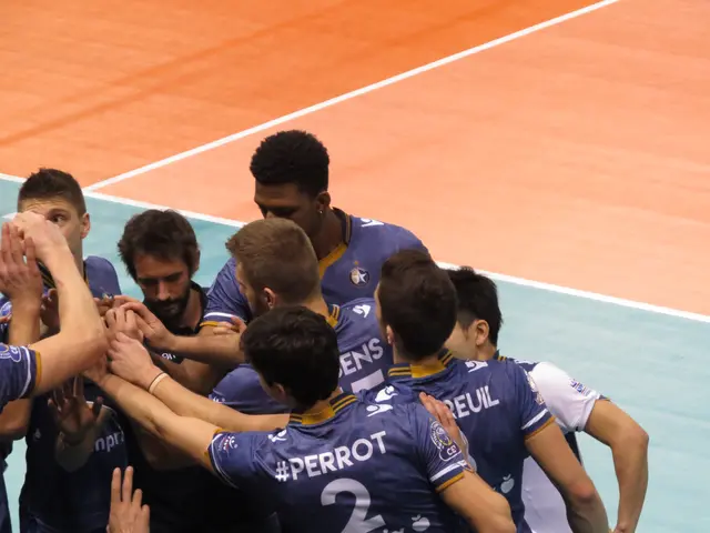 The image shows a group of men standing on top of a volleyball court, all wearing blue t-shirts...