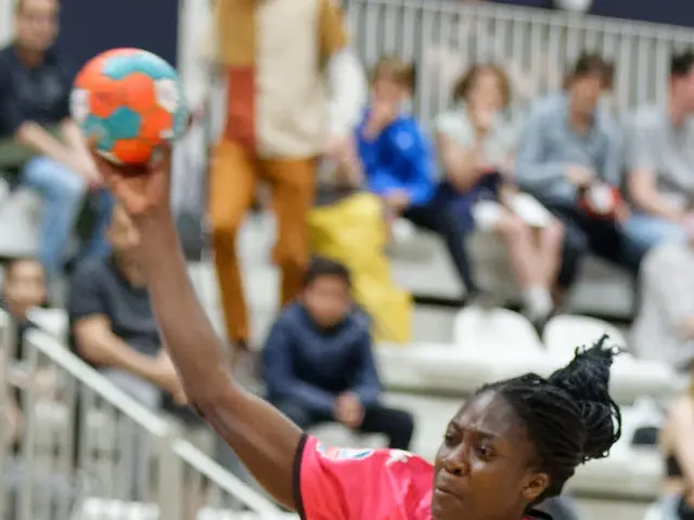 The image shows two women playing a game of handball in front of a crowd of people sitting on...