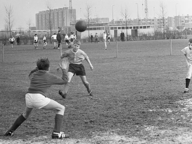 The image shows a group of young boys playing soccer on a field surrounded by buildings, trees,...