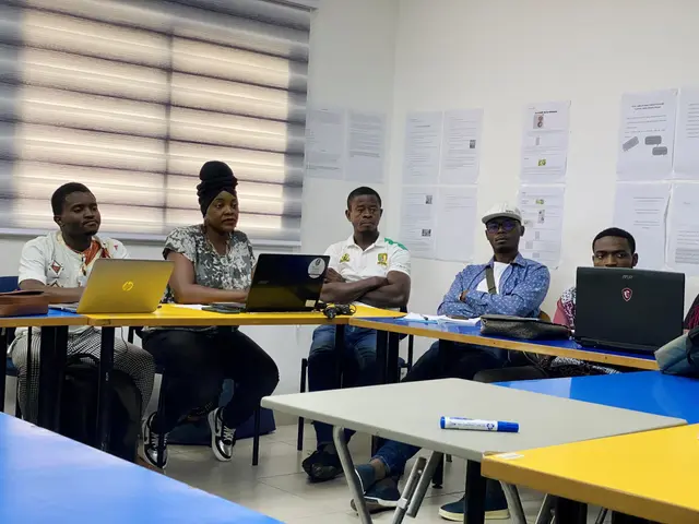 The image shows a group of people sitting around a table with laptops, bags, pens, and other...