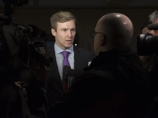 The image shows a man in a suit and tie talking to reporters, with two people holding cameras on...