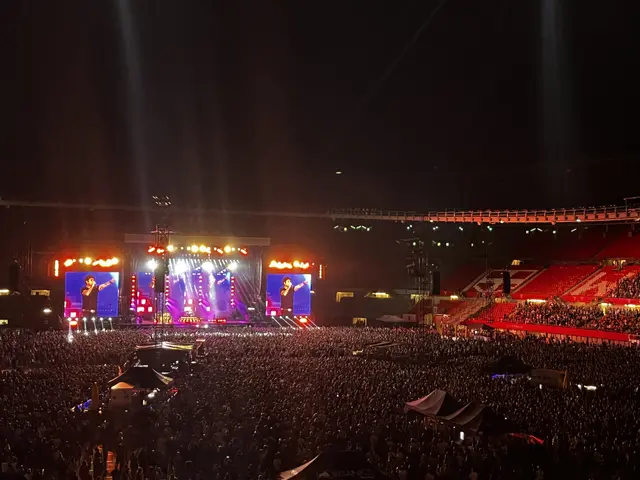 The image shows a large crowd of people gathered at a concert, with a stage in the center of the...