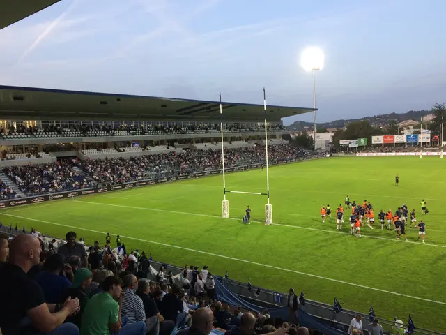 The image shows a large crowd of people watching a rugby match in a stadium. The stadium is filled...