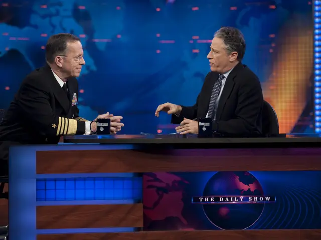 The image shows two men sitting at a table in front of a TV screen, both wearing black blazers,...