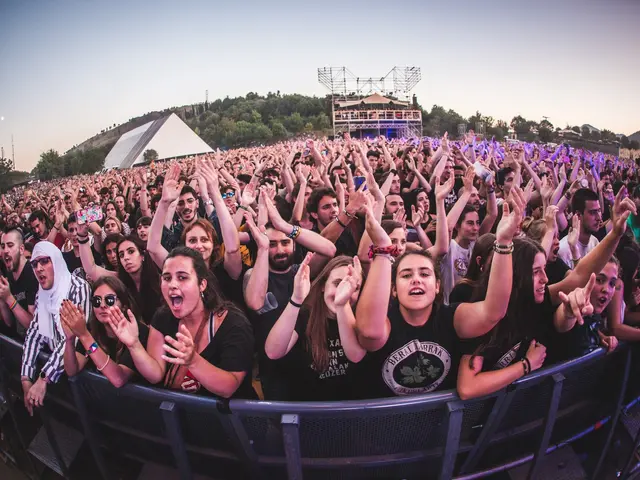 The image shows a large crowd of people at a music festival with their hands in the air, standing...