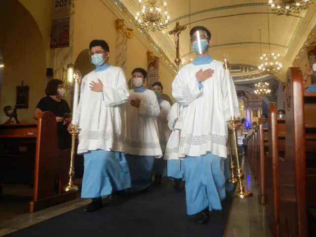 The image shows a group of people wearing masks walking down a church aisle, with benches on either...