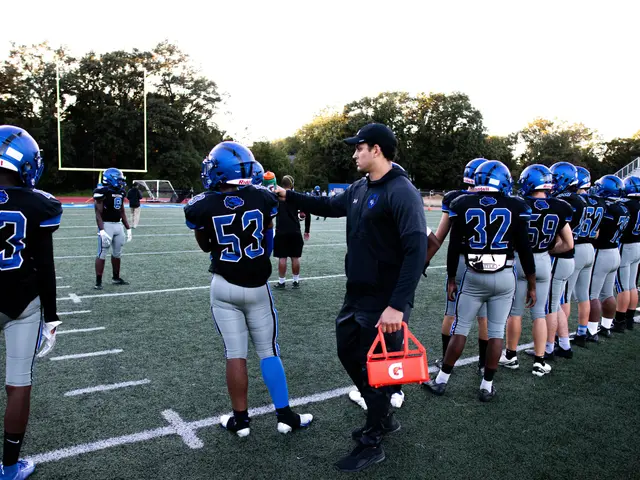 The image shows a football coach talking to his team on the field. He is wearing a cap and holding...