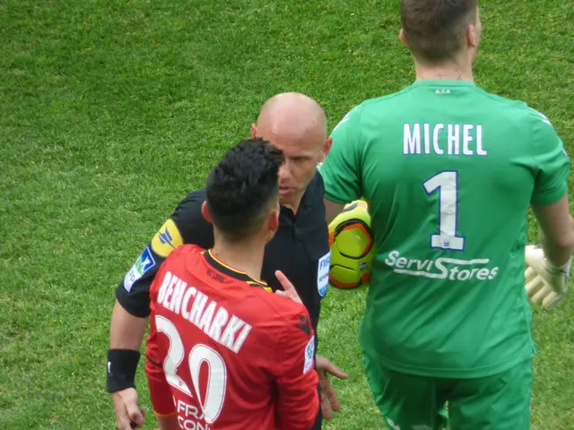The image shows a referee talking to two soccer players on a field, with one of them holding a ball...