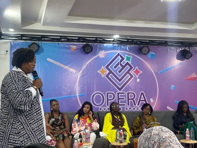 The image shows a woman standing in front of a group of women sitting on a couch, holding a...