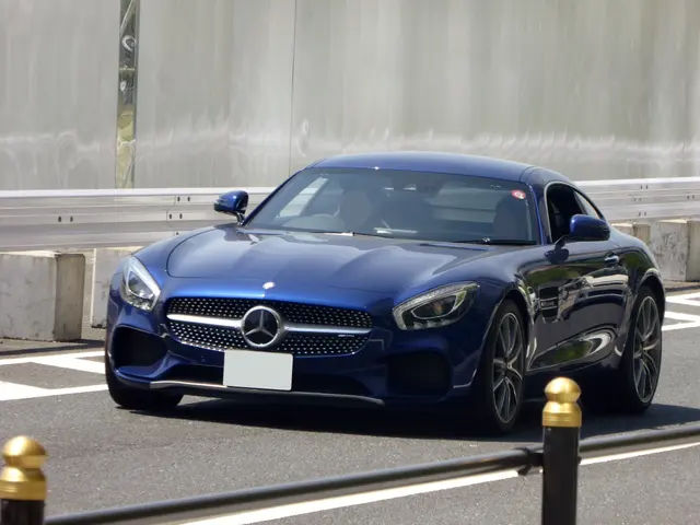 The image shows a blue Mercedes-Benz AMG GT Coupe driving down a street, with a wall in the...