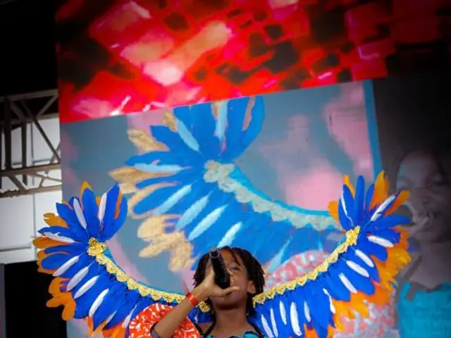 The image shows a woman in a blue and orange outfit standing on a stage, holding a microphone and...