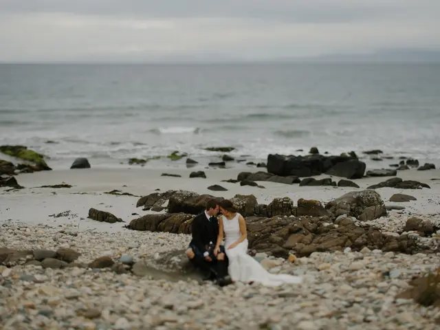 The image shows a bride and groom sitting on a rocky beach, with the ocean in the background and...