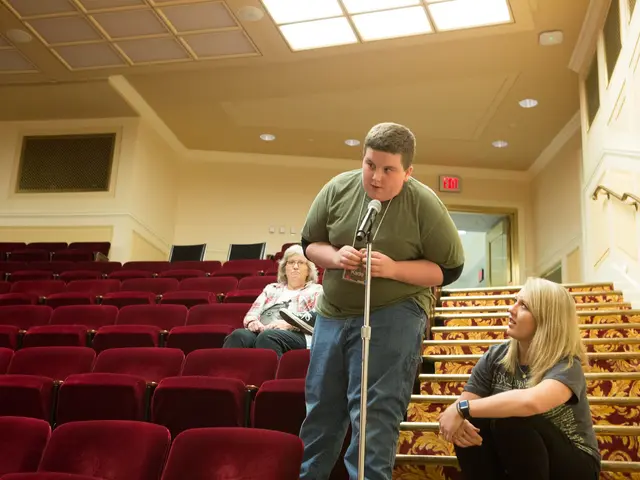 The image shows a man standing in front of a crowd of people in a theater. He is wearing a green...