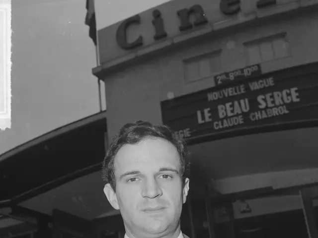 The image shows a man in a suit and tie standing in front of a cinema, with a building in the...