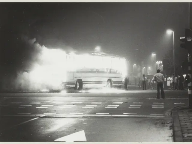 The image shows a black and white photo of a bus engulfed in flames on the side of the road, with a...