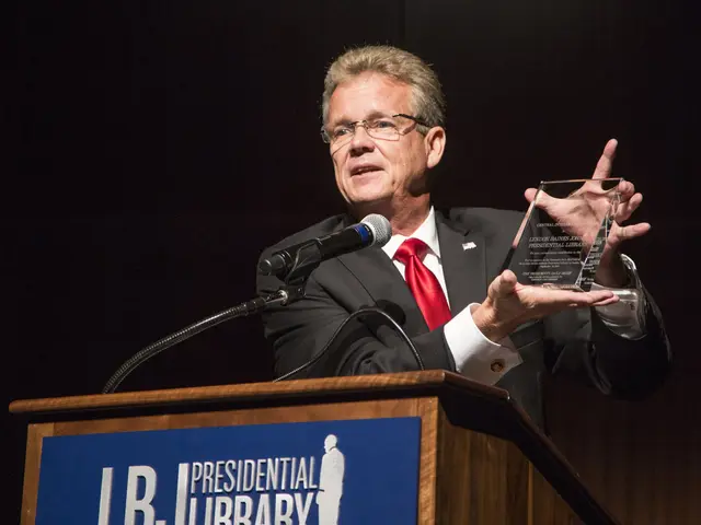 The image shows a man in a suit and tie standing at a podium, holding an award in his hand. He is...