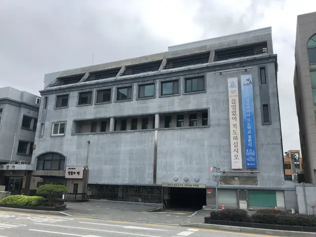 The image shows a large building with a lot of windows on the side of it, surrounded by plants, a...