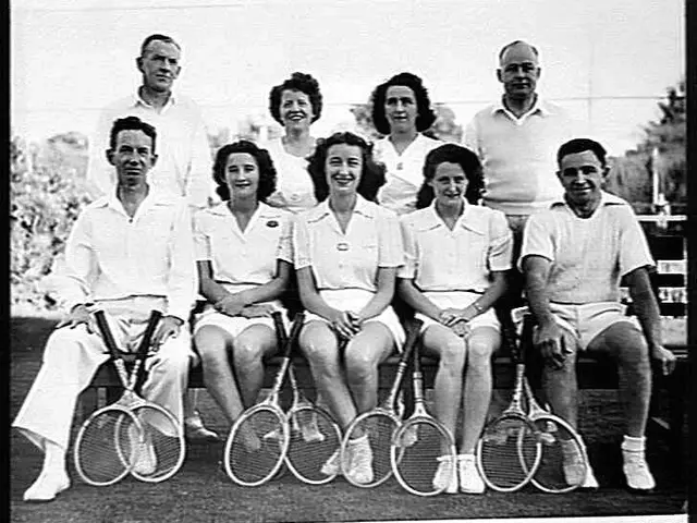 The image shows a black and white photo of a group of people sitting on a bench, smiling and...