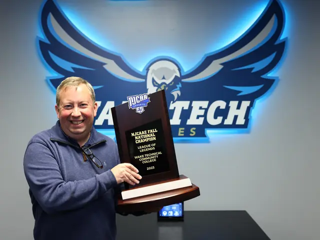 The image shows a man standing and smiling while holding a plaque in front of a wall with a logo...