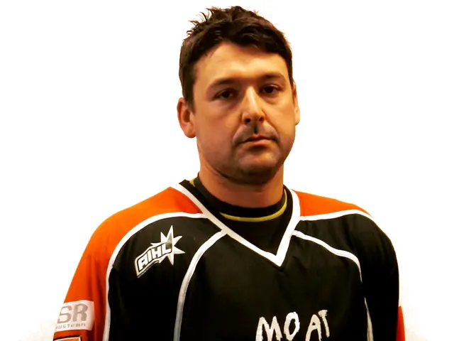 The image shows a man in a black and orange hockey jersey standing against a white background. He...