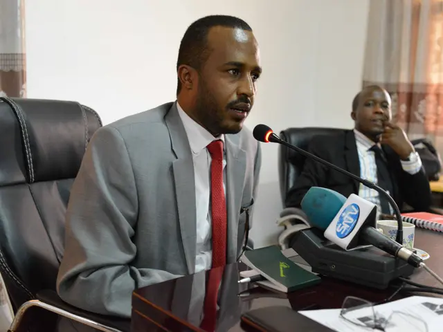 The image shows two men sitting at a table with microphones in front of them. The man on the left...
