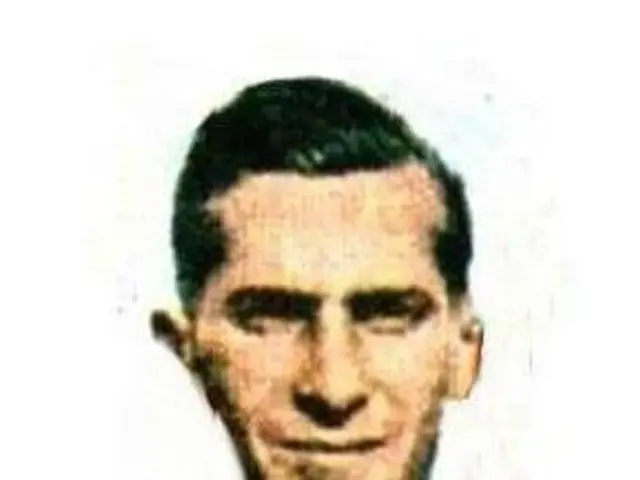 The image shows a man with a beard wearing a red and white shirt, smiling against a white...