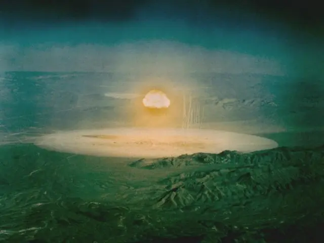 The image shows an atomic bomb exploding over a large crater in the middle of a desert, with hills...
