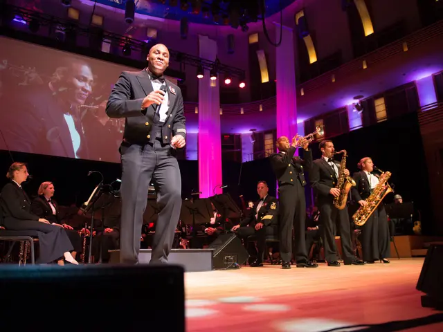 The image shows a man in a suit and tie standing on a stage with a microphone in his hand,...